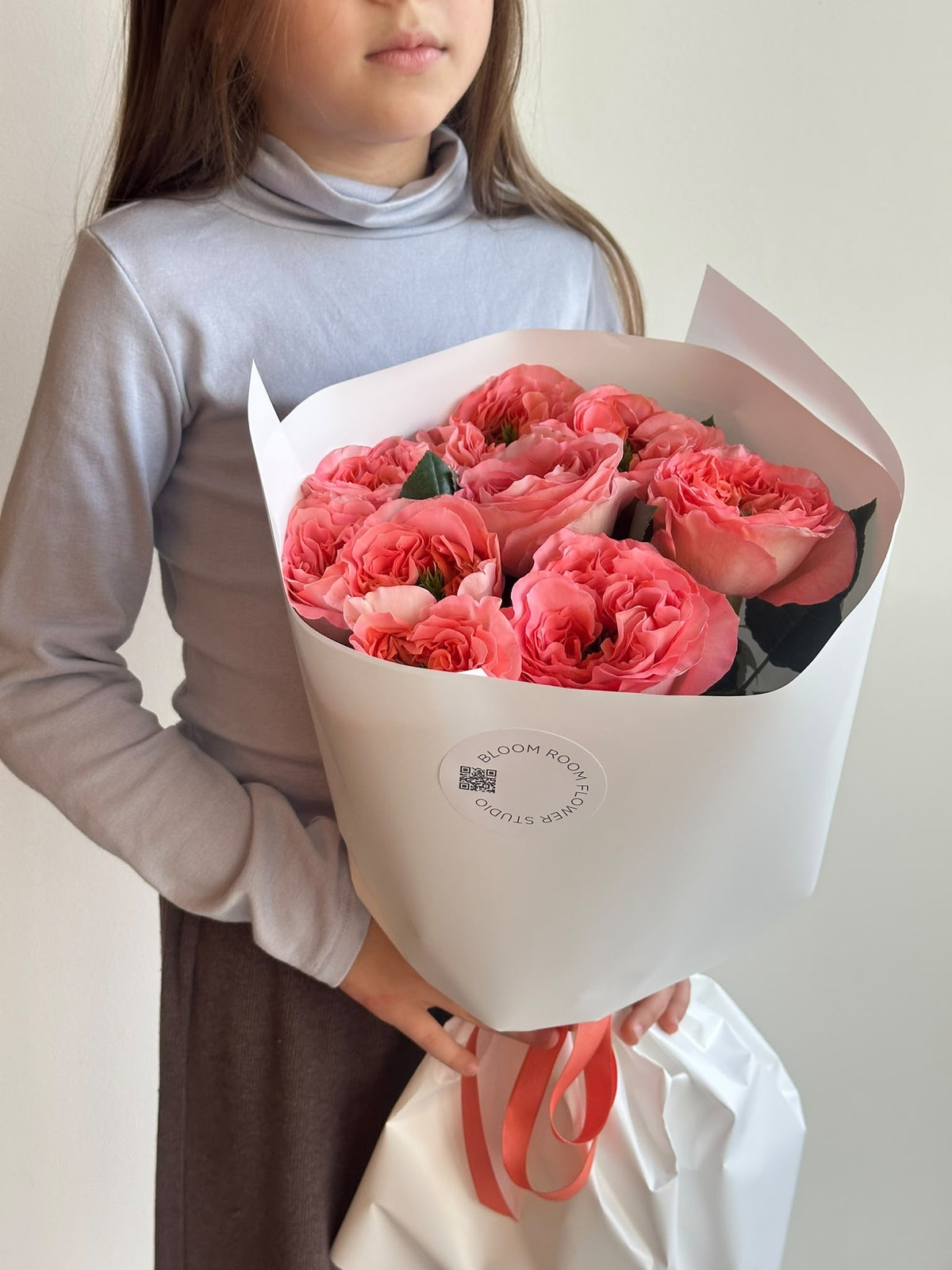 „Coral Kiss“ kimp korallroosadest pojengikujulistest roosidest valges pakendis
