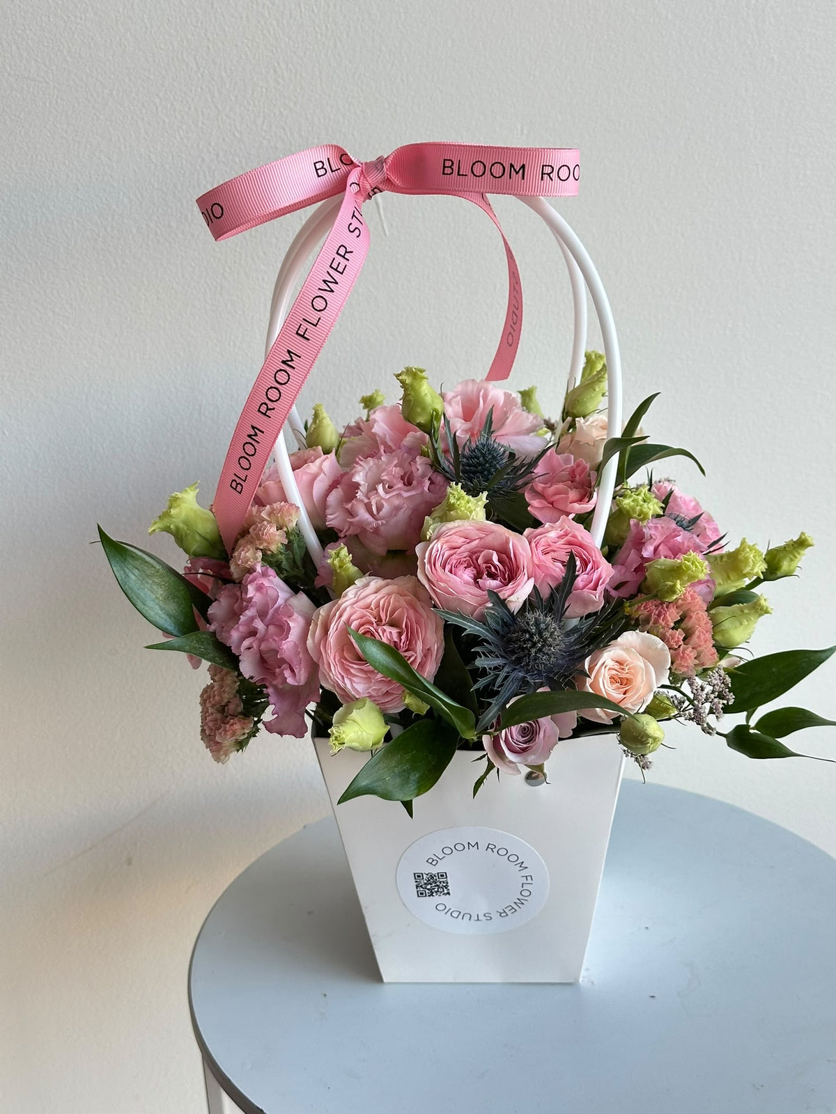 “Petite Romance” mini flower bag with pink spray roses, carnations, eustoma and eryngium in a white Bloom Room bag